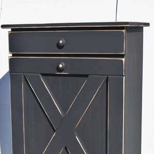 May include: Black wooden trash can with a farmhouse style X-shaped door and two drawers. The can has a distressed finish and white accents.
