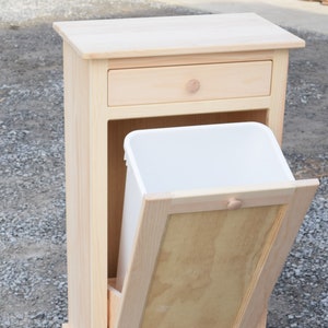 May include: A wooden trash can cabinet with a single drawer and a hinged door. The cabinet is made of unfinished pine wood and has a white plastic trash can inside.