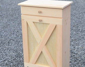May include: A wooden trash can with a farmhouse style door and a drawer. The can is made of unfinished pine wood and has black hinges. The door has a diagonal X design.
