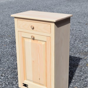 May include: A light wood trash can cabinet with a single drawer and a door with black metal handles. The cabinet is standing on a gravel surface.