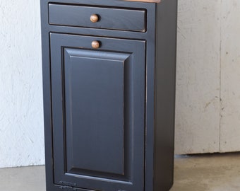 May include: A black painted wooden trash can with a single drawer and a hinged door. The door has a raised panel design and the drawer has a knob. The top of the can is a light brown wood.