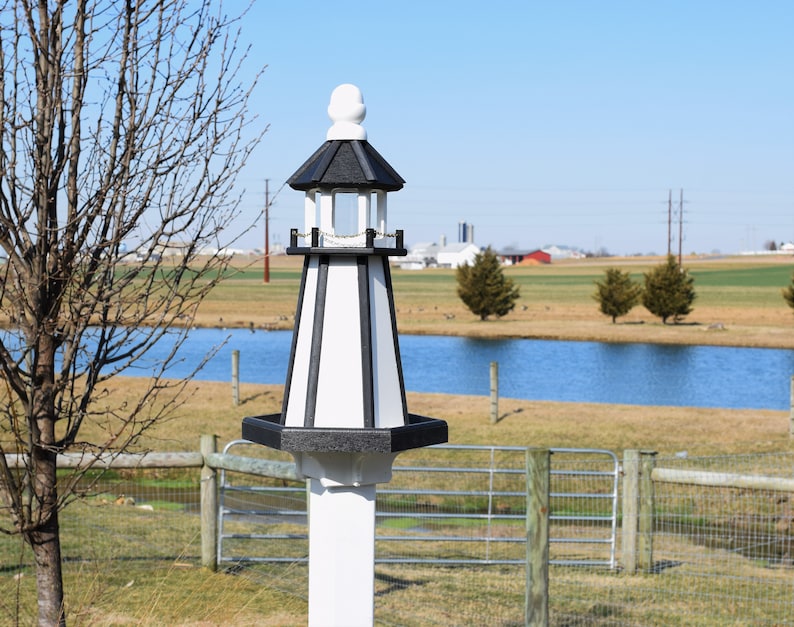 May include: A black and white birdhouse shaped like a lighthouse with a white post. The birdhouse has a black roof and a white base. It is sitting in a field with a pond in the background.