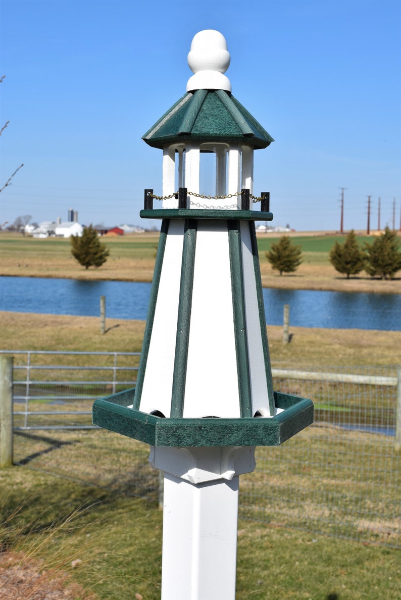 May include: A white and green birdhouse shaped like a lighthouse with a small, white, rounded roof. The birdhouse has a green base and a white post.