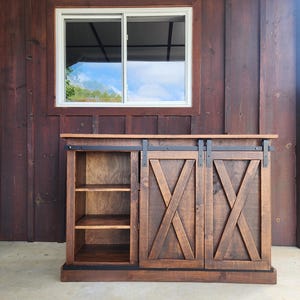 May include: A rustic wooden cabinet with two sliding barn doors. The doors have a criss-cross pattern and black hardware. The cabinet has three shelves on the left side.