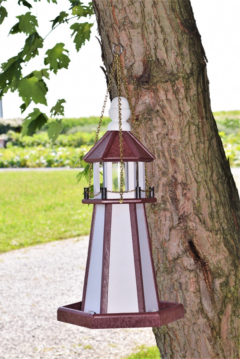 May include: A white and burgundy bird feeder shaped like a lighthouse with a gold chain hanging from a tree. The feeder has a clear plastic window and a hexagonal base.