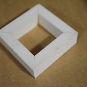 May include: A white, geometric, diamond-shaped object. The object is made of a solid material and has a square opening in the center. The edges are beveled, and the object is sitting on a brown surface.