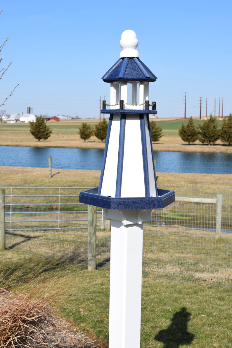 May include: A white and blue birdhouse shaped like a lighthouse with a white post. The birdhouse has a blue roof and a white base. The birdhouse is in a yard with a pond and a fence in the background.