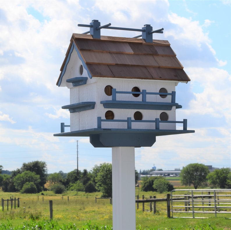 以下が含まれることがあります： 茶色の屋根瓦のついた白と青の鳥小屋。鳥小屋は複数の巣箱があり、白い柱に取り付けられています。