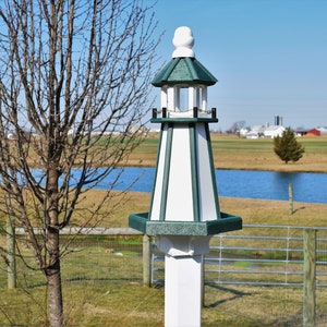 May include: A white and green birdhouse shaped like a lighthouse with a green roof and a white base. The birdhouse is standing in a field with a pond in the background.