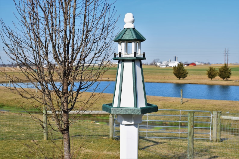 May include: A white and green birdhouse shaped like a lighthouse with a green roof and a white base. The birdhouse is standing in a field with a pond in the background.
