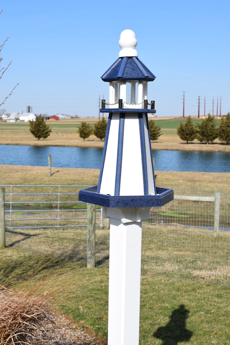 May include: A white and blue birdhouse shaped like a lighthouse with a white post. The birdhouse has a blue roof and a white base with blue trim. The birdhouse is in a field with a pond in the background.