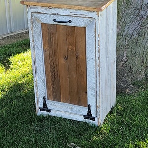 May include: White painted wooden trash can with a brown wood door and black hinges. The can has a black handle and sits on a green grassy lawn.