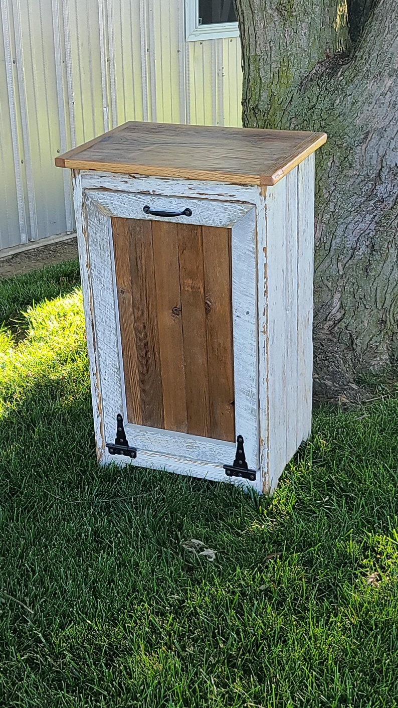 May include: White painted wooden trash can with a brown wood door and black hinges. The can has a black handle and sits on a green grassy lawn.