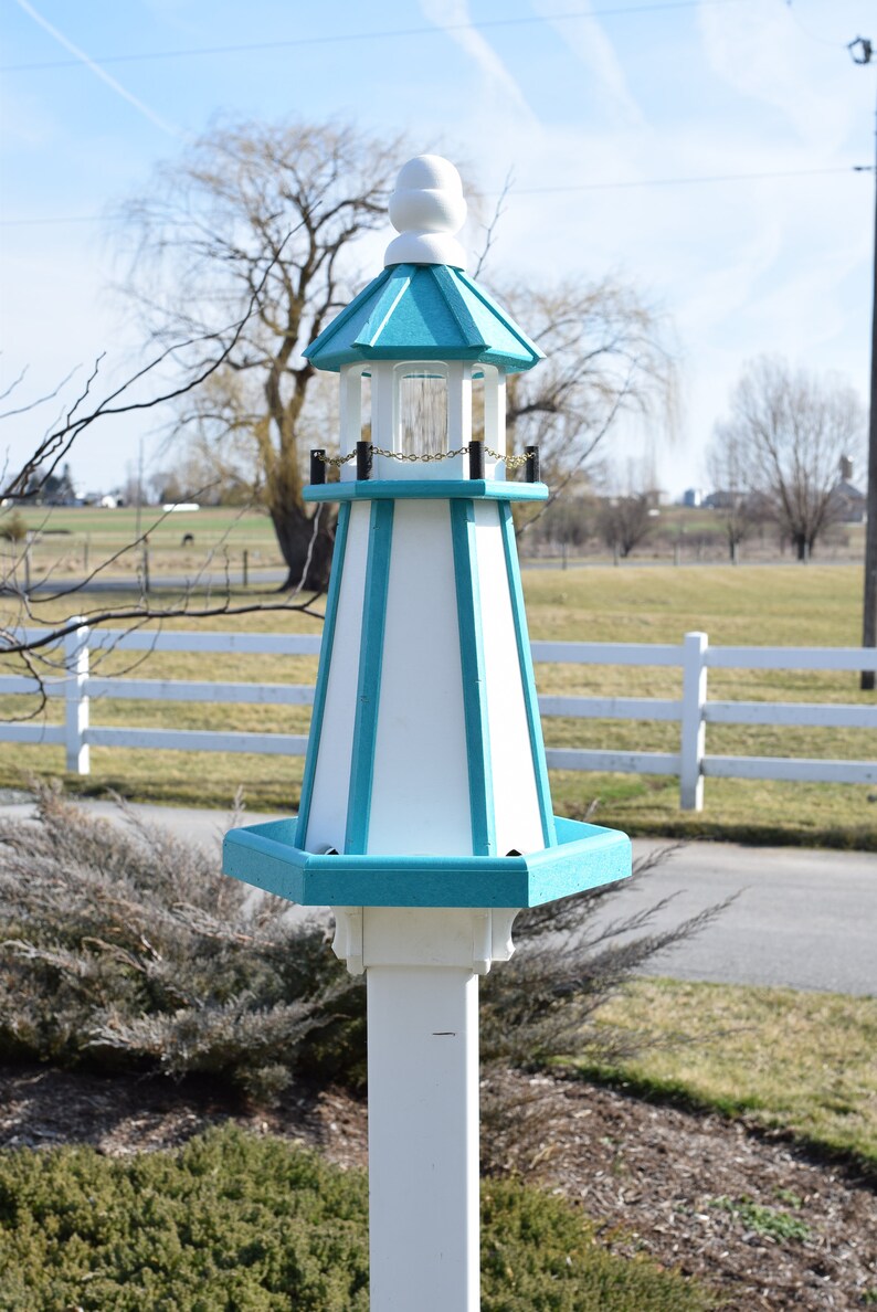 May include: A white and teal blue lighthouse birdhouse with a gold chain hanging from the top. The birdhouse is mounted on a white wooden pole.