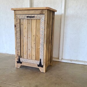 May include: A rustic wooden trash can with a single door and black metal hinges. The can is made of reclaimed wood and has a natural wood finish.