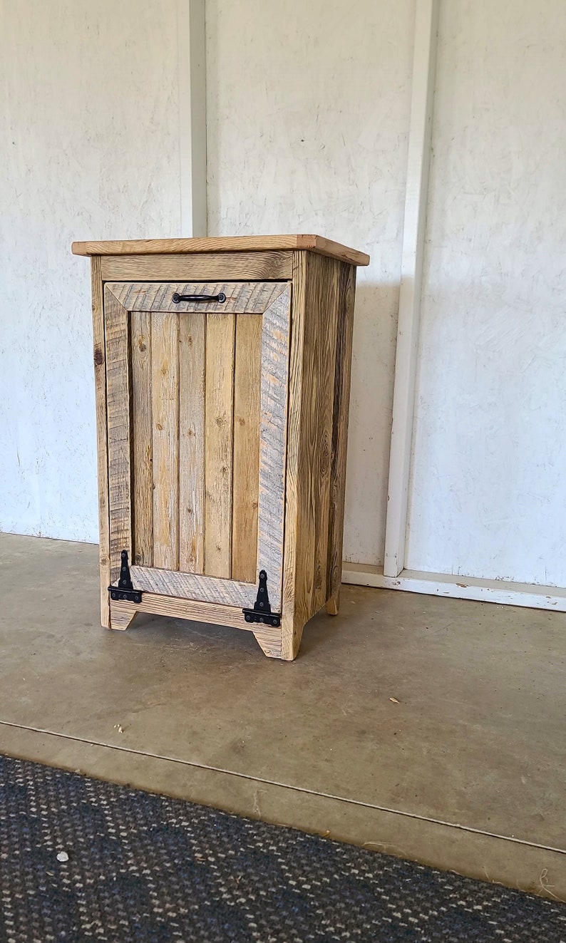 May include: A rustic wooden trash can with a single door and black metal hinges. The can is made of reclaimed wood and has a natural wood finish.