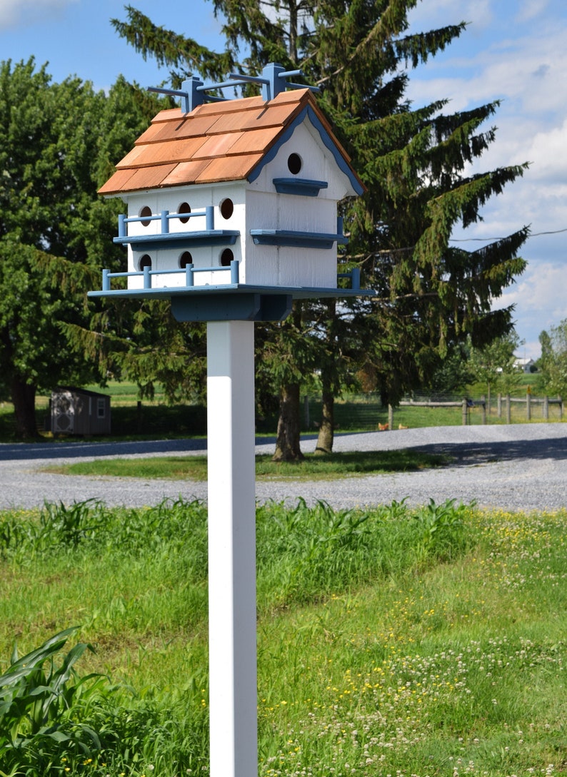 以下が含まれることがあります： 白い柱の上に置かれた、白と青の鳥小屋。茶色の屋根と、複数の巣箱と装飾的なデザインが施されています。
