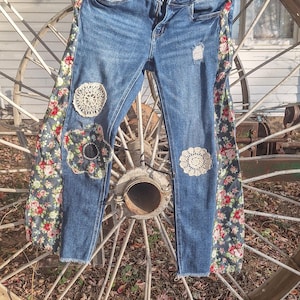 May include: Blue denim jeans featuring side panels with a floral pattern of red and pink roses on a dark background. The jeans include white circular crochet lace appliques, a distressed patch on the left leg, and a raw, frayed hemline.