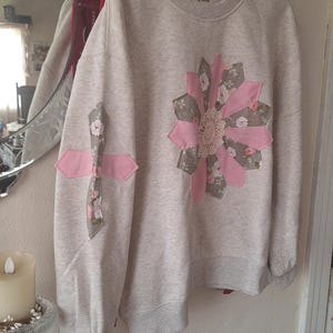 May include: A light heather gray crewneck sweatshirt featuring a large, multi-petaled flower appliqué on the chest made of pink fabric and floral-patterned fabric with a central lace doily. A matching cross-shaped appliqué is on the sleeve.