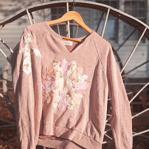 May include: A dusty rose long-sleeve sweatshirt featuring a large, multi-colored floral fabric flower appliqué on the center chest and a smaller cross-shaped floral fabric appliqué on the right sleeve, with a V-neckline and ribbed cuffs.