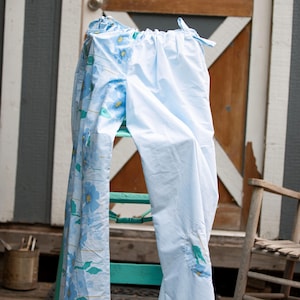 Può includere: Un paio di pantaloni azzurri caratterizzati da un motivo floreale su una gamba e da un tessuto azzurro a tinta unita sull'altra. I pantaloni presentano un elastico in vita con chiusure a laccio e un piccolo dettaglio a toppa floreale.