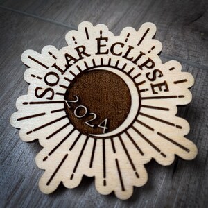 Könnte beinhalten: Ein Holz-Anhänger mit einem Sonnenstrahlen-Design und dem Text "Solar Eclipse 2024".