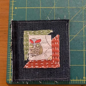 May include: A square denim fabric patch featuring a central white square with a cartoon owl illustration, layered over green and orange textured fabric scraps, all secured with white hand-stitched running stitches on a dark blue denim base.