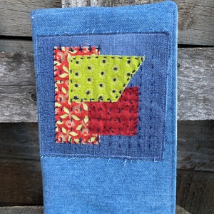 May include: A blue denim notebook cover with a patchwork design featuring red, green, and yellow fabric squares. The squares are sewn together with a contrasting thread.