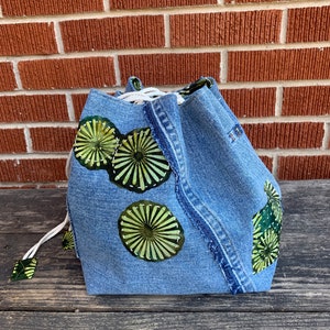 May include: A blue denim drawstring bag with green and yellow circular designs. The bag is made from recycled denim and has a unique, handmade look.