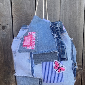 Może przedstawiać: Niebieska torba z denimu wykonana z odzyskanego materiału dżinsowego. Torba ma patchworkowy wzór z różnymi odcieniami niebieskiego denimu i różową aplikacją motyla. Torba ma biały sznurek.