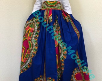 May include: A long, blue maxi skirt with a colorful African print pattern. The skirt has a high waist and is pleated. The skirt is paired with a white button-down shirt.