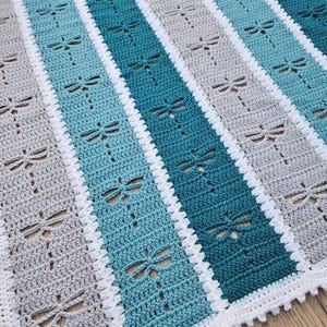 May include: Hand-crocheted blanket with a dragonfly design. The blanket has alternating stripes of grey and teal, with a white border. The dragonfly cutouts add a decorative touch to the handmade blanket.