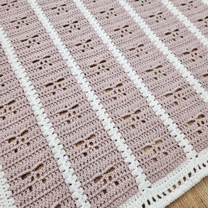 May include: A handmade crochet blanket in a soft pink hue with white stripes. The blanket features a repeating pattern of small, openwork designs. The edges are finished with a white border, creating a cozy and textured appearance.