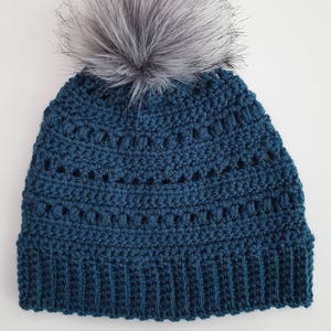May include: A teal blue crochet beanie with a gray faux fur pom-pom on top. The hat features a ribbed brim and a textured stitch pattern. This handmade winter hat is a cozy accessory.