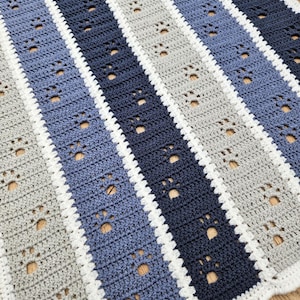 May include: A handmade crochet blanket featuring a striped pattern in shades of blue and gray, with white borders. The design incorporates paw print cutouts throughout the blanket. The blanket is a cozy, decorative item.