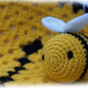 Puede incluir: Un juguete de abeja de crochet con rayas negras y amarillas y alas blancas. La abeja está sentada en una manta de crochet amarilla.