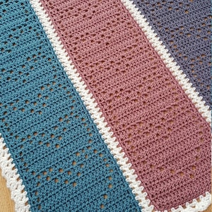 Può includere: Una coperta all'uncinetto con tre strisce di colori diversi: turchese, rosa e grigio. La coperta ha un bordo all'uncinetto bianco.