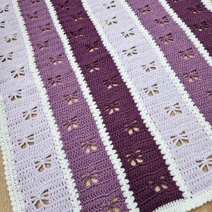 May include: A handmade crochet blanket with a striped design in shades of purple and white. The blanket features a repeating dragonfly pattern and a white scalloped border. The colour palette includes light lavender, medium purple, and deep violet.