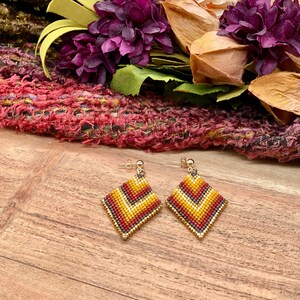 May include: A pair of gold tone earrings with a geometric diamond shape design. The earrings are made of small beads in shades of red, orange, yellow, and black.