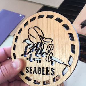 May include: A wooden coaster featuring a black and white illustration of a cartoon bee holding a wrench. The text "SEABEES" is printed below the bee.