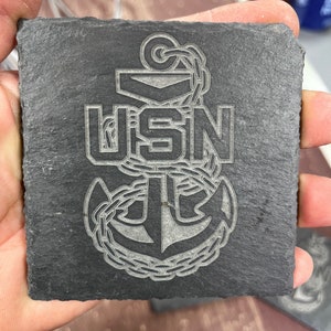 May include: A black slate coaster with a white engraved image of an anchor and the letters "USN".