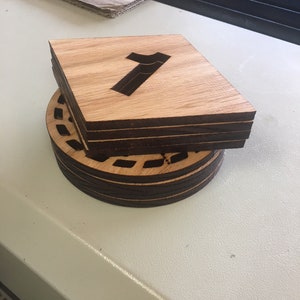 May include: A stack of four wooden coasters with a laser-cut design. The top coaster features the number "7" cut out of the centre. The coasters are made of light brown wood with a dark brown stain around the edges.