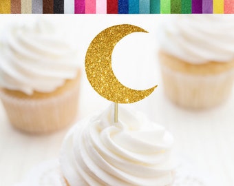 May include: Gold glitter crescent moon cupcake topper. The topper is on a white cupcake with white frosting. The cupcake is on a white doily. The background is white.