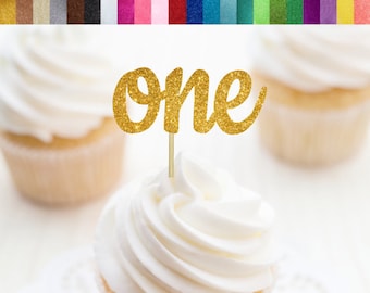 May include: Gold glitter cupcake topper with the word "one" in a cursive font. The topper is on a white cupcake with white frosting.