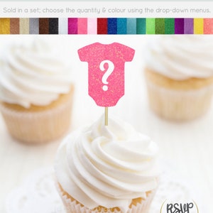May include: Pink glitter cupcake topper shaped like a baby onesie with a question mark. The topper is on a stick and is ready to be placed on a cupcake.