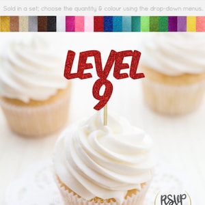 Pode incluir: Um cupcake com cobertura branca e um palito vermelho brilhante "LEVEL 9". O cupcake está em uma toalha de mesa branca, com dois outros cupcakes desfocados ao fundo. A imagem inclui o texto "Cupcake Toppers/Food Picks - Glitter Cardstock".