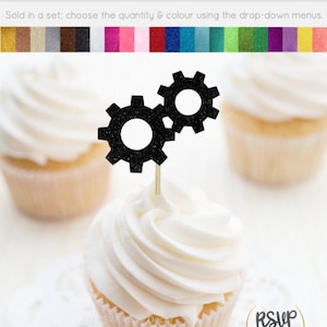 以下が含まれることがあります： 黒のグリッターギアのカップケーキトッパー2つ。ギアは互いに噛み合っており、つまようじに取り付けられています。カップケーキトッパーは、白いフロスティングをかけた白いカップケーキの上にあります。