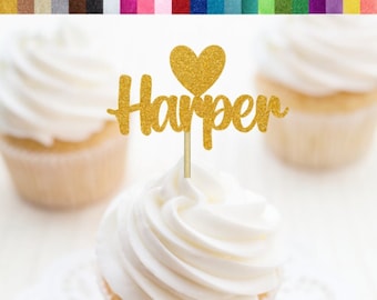 Op de afbeelding: Gouden glitter cupcake topper met de naam "Harper" en een hart. De topper is op een witte cupcake met witte frosting.