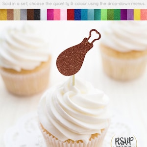 May include: A cupcake topped with a glitter-covered brown turkey leg cupcake topper. The cupcake topper is made of cardstock and has a gold glitter finish. The cupcake is frosted with white frosting. The cupcake topper is sold in a set and can be customized with different colors.