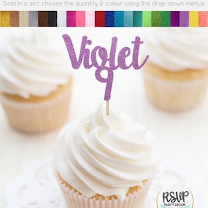 Op de afbeelding: Paarse glitter cupcake topper met de naam "Violet" en het nummer "9" erop. De topper zit op een cupcake met witte frosting.